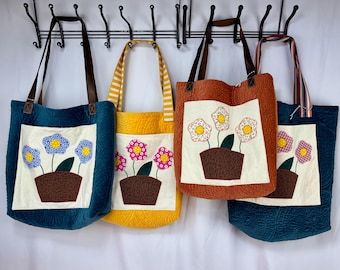 Könnte beinhalten: Vier gesteppte Tragetaschen mit unterschiedlichen Stofffarben und Blumendesigns. Die Taschen hängen an einem schwarzen Metallgestell. Jede Tasche hat ein anderes farbiges Blumendesign mit einem braunen Topf.