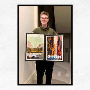 May include: A person holding two framed posters. The poster on the left is a vintage-style illustration of Yosemite National Park with the text "Yosemite National Park". The poster on the right is a vintage-style illustration of Zion National Park with the text "Zion National Park".