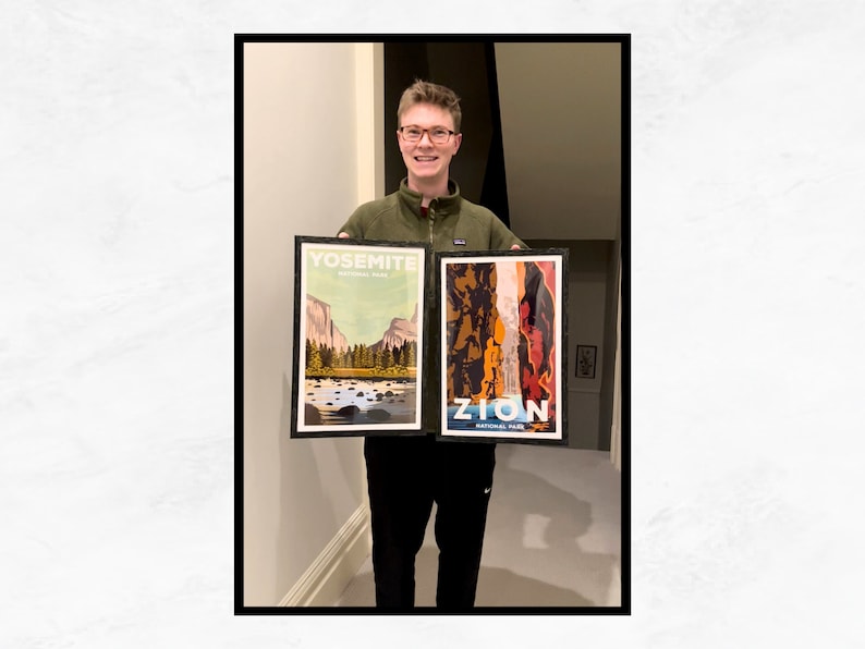 May include: A person holding two framed posters. The poster on the left is a vintage-style illustration of Yosemite National Park with the text "Yosemite National Park". The poster on the right is a vintage-style illustration of Zion National Park with the text "Zion National Park".