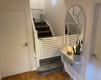 May include: White wooden baby gate with a white railing and grey carpet stairs in the background. A white console table with a plant and a candle on top is in the foreground.