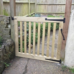 May include: A light-coloured wooden garden gate with vertical slats and a rectangular frame. The gate features black hinges and a latch. It is set in a garden with a stone wall and a wooden fence in the background.