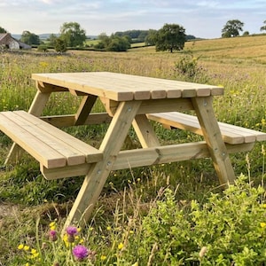May include: A light-colored wooden picnic table featuring a rectangular tabletop and two attached benches on opposite sides. The structure is supported by angled legs and cross-bracing, constructed from horizontal wooden slats with visible metal bolts.