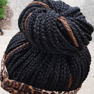 Pu&ograve; includere: Capelli neri intrecciati acconciati a chignon, con una treccia marrone di accento. Una fascia con un motivo leopardato marrone e beige circonda la base dei capelli. I capelli sono acconciati su una testa di manichino bianca.