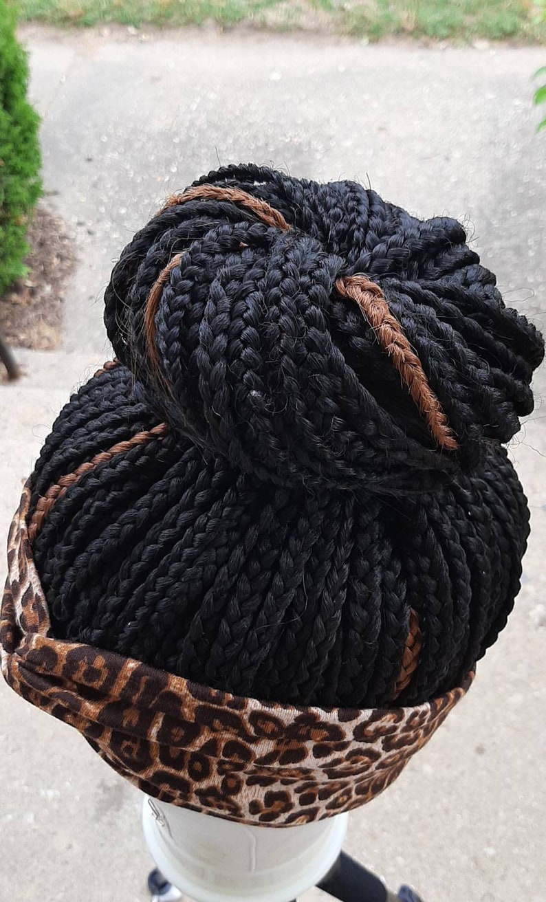 Pu&ograve; includere: Capelli neri intrecciati acconciati a chignon, con una treccia marrone di accento. Una fascia con un motivo leopardato marrone e beige circonda la base dei capelli. I capelli sono acconciati su una testa di manichino bianca.