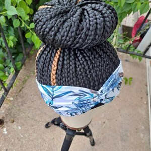 Pu&ograve; includere: Una parrucca nera intrecciata acconciata a chignon, con un accento di treccia marrone. La parrucca &egrave; indossata su una testa di manichino, con una fascia floreale bianca e blu. Il manichino &egrave; montato su un treppiede nero.