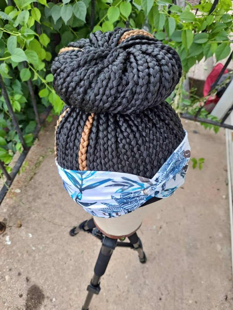 Pu&ograve; includere: Una parrucca nera intrecciata acconciata a chignon, con un accento di treccia marrone. La parrucca &egrave; indossata su una testa di manichino, con una fascia floreale bianca e blu. Il manichino &egrave; montato su un treppiede nero.