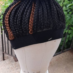 Pu&ograve; includere: Una parrucca intrecciata nera con riflessi marroni, acconciata a chignon, &egrave; esposta su una testa di manichino. La testa &egrave; montata su un treppiede. Lo sfondo comprende vegetazione e una ringhiera metallica.