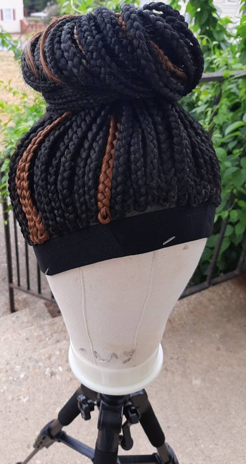 Pu&ograve; includere: Una parrucca intrecciata nera con riflessi marroni, acconciata a chignon, &egrave; esposta su una testa di manichino. La testa &egrave; montata su un treppiede. Lo sfondo comprende vegetazione e una ringhiera metallica.