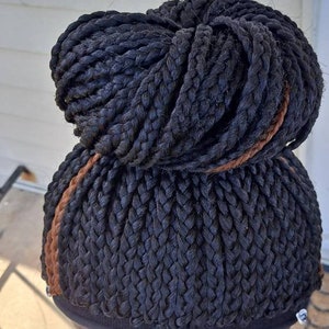 Pu&ograve; includere: Parrucca nera intrecciata a chignon. La parrucca presenta un'evidenziazione marrone scuro ed &egrave; montata su un supporto nero. Le trecce sono fitte, creando un aspetto strutturato. Il supporto per parrucca ha dettagli argentati.