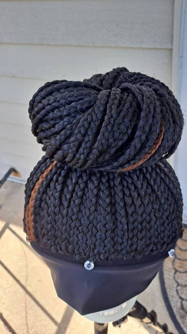 Pu&ograve; includere: Parrucca nera intrecciata a chignon. La parrucca presenta un'evidenziazione marrone scuro ed &egrave; montata su un supporto nero. Le trecce sono fitte, creando un aspetto strutturato. Il supporto per parrucca ha dettagli argentati.