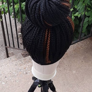 Pu&ograve; includere: Una parrucca nera intrecciata acconciata a chignon con riflessi marroni. La parrucca &egrave; esposta su una testa di manichino bianca, montata su un treppiede nero. Lo sfondo presenta fogliame verde e una ringhiera in metallo nero.