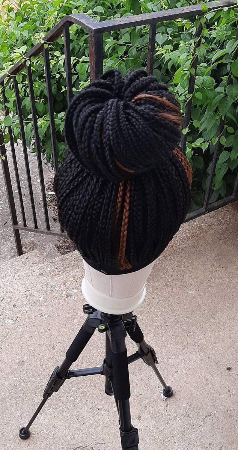 Pu&ograve; includere: Una parrucca nera intrecciata acconciata a chignon con riflessi marroni. La parrucca &egrave; esposta su una testa di manichino bianca, montata su un treppiede nero. Lo sfondo presenta fogliame verde e una ringhiera in metallo nero.
