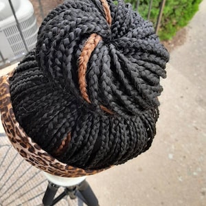 Pu&ograve; includere: Parrucca intrecciata nera con un accento marrone, acconciata a chignon. La parrucca &egrave; posta su una testa di manichino bianca, con un tessuto leopardato intorno alla base. La parrucca &egrave; su un treppiede nero.