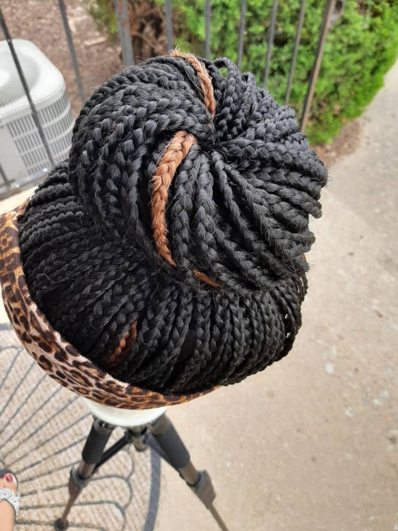 Pu&ograve; includere: Parrucca intrecciata nera con un accento marrone, acconciata a chignon. La parrucca &egrave; posta su una testa di manichino bianca, con un tessuto leopardato intorno alla base. La parrucca &egrave; su un treppiede nero.