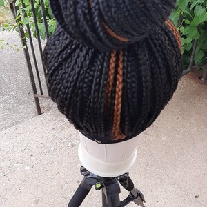 Pu&ograve; includere: Una parrucca nera intrecciata acconciata in uno chignon alto con riflessi ramati. La parrucca &egrave; esposta su una testa di manichino bianca, montata su un treppiede nero. Lo sfondo comprende vegetazione e una ringhiera metallica.