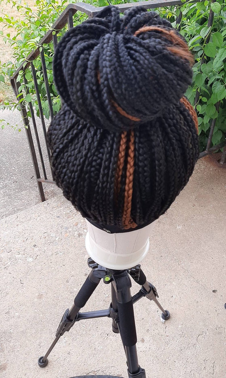 Pu&ograve; includere: Una parrucca nera intrecciata acconciata in uno chignon alto con riflessi ramati. La parrucca &egrave; esposta su una testa di manichino bianca, montata su un treppiede nero. Lo sfondo comprende vegetazione e una ringhiera metallica.