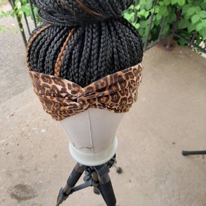 Pu&ograve; includere: Una parrucca intrecciata nera, acconciata a chignon alto, con ciocche marroni. La parrucca &egrave; adornata da una fascia con stampa leopardata. La parrucca &egrave; esposta su una testa di manichino, montata su un treppiede nero.