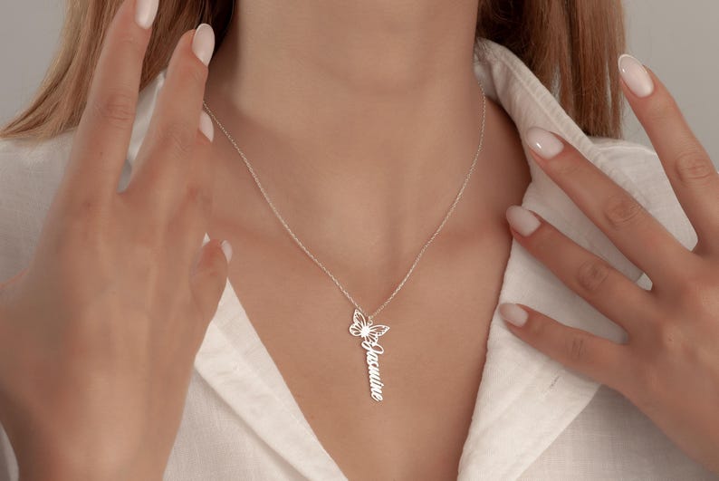 May include: A silver necklace featuring a butterfly pendant and the name "Jasmine" engraved vertically. The butterfly has a sunburst design. The necklace is worn over a white shirt.
