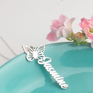 May include: A silver pendant necklace with a butterfly design and the name "Jasmine" in cursive. The necklace is displayed on a teal surface, with pink flowers in the background. The necklace is a beautiful piece of jewellery.