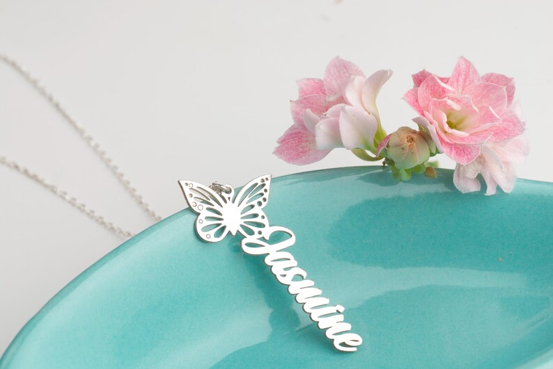 May include: A silver pendant necklace with a butterfly design and the name "Jasmine" in cursive. The necklace is displayed on a teal surface, with pink flowers in the background. The necklace is a beautiful piece of jewellery.