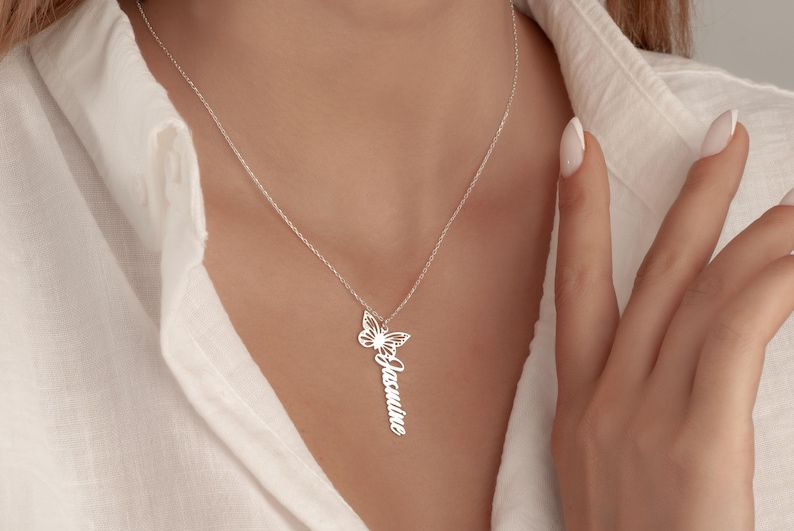 May include: A silver necklace with a butterfly pendant and a vertical nameplate. The butterfly design is detailed, and the name "Jasmine" is inscribed. The necklace is shown against a white shirt.