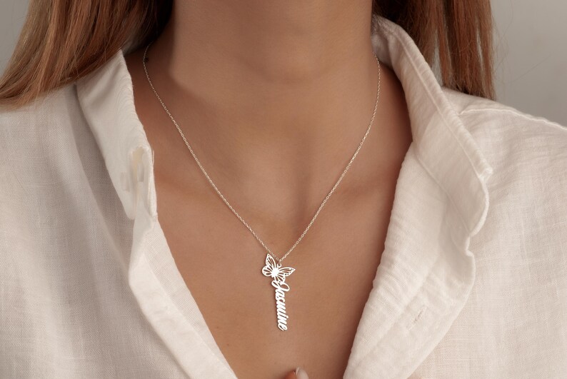 May include: A silver necklace with a butterfly pendant and a vertical nameplate reading "Jasmine". The necklace is worn against a white shirt.