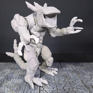 May include: A gray, sculpted monster figurine with a large open mouth, sharp claws, and a segmented tail. The creature has a textured, rough surface and is posed on a stone-like surface. The figurine is a detailed representation of a fantasy creature.