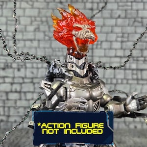 May include: A silver and gray action figure head with a fiery orange head and a skeletal face. The figure has a metallic chest and arms. Chains hang from the head. A blue sign at the bottom reads "*ACTION FIGURE NOT INCLUDED."