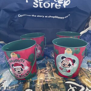 Op de afbeelding: Vier kleine rode metalen emmers met groene handvatten. Elke emmer heeft een ander kerstthema met Mickey Mouse en Minnie Mouse. De emmers zeggen "Naughty or Nice?" en "Mickey's Christmas".