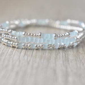 May include: A multi-strand bracelet featuring alternating segments of small, pale blue translucent beads and metallic silver-toned beads. The design consists of three distinct rows of beads arranged in a circular pattern, creating a layered appearance.