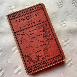 May include: A vintage travel guide for Torquay and South Devon, published by Ward, Lock & Co. The cover is a reddish-orange with a black map and text. It is part of the "Illustrated Guide Books" series.
