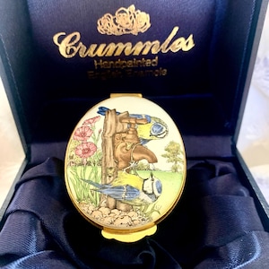 May include: An oval Crummles handpainted English enamel box featuring a scene of two blue tits perched on a garden water tap. The lid displays pink flowers, green grass, and a tree. The box is presented in a dark blue satin-lined case with gold text.