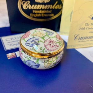 May include: A round, hand-painted enamel trinket box with a gold-tone metal rim. The exterior features a cream-coloured background decorated with a floral pattern of pink, blue, and yellow sweet pea flowers with green vines and curling tendrils.