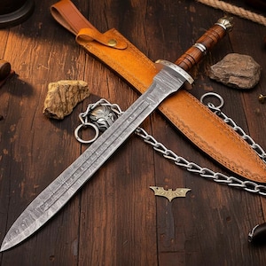 May include: A detailed image of a sword with a patterned blade and a wooden handle. The sword rests in a tan leather sheath, with a matching leather strap. A metal chain is attached to the sheath. The background is a dark wooden surface.