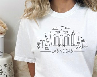 May include: White t-shirt featuring a black outline of the Las Vegas skyline, including the Eiffel Tower, the Bellagio fountain, and the Stratosphere tower. The text "LAS VEGAS" is printed below the skyline.
