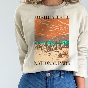 May include: A beige sweatshirt with a vintage-style graphic of Joshua Tree National Park. The graphic features a desert landscape with Joshua trees, mountains, and a sunset sky.