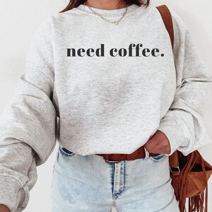 May include: A grey sweatshirt with the text "need coffee." printed in black on the front. The sweatshirt is being worn by a person with a brown leather bag slung over their shoulder.