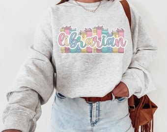 Peut inclure: Un sweat-shirt gris avec un motif de livres colorés et le mot "bibliothécaire" dans une police de caractères fantaisiste.