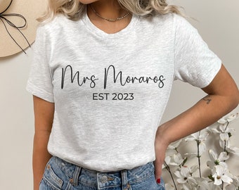 Puede incluir: Una camiseta gris claro con el texto "Mrs. Moraros EST 2023" en escritura negra. La camiseta es de cuello redondo y está hecha de un material suave. La persona lleva vaqueros azules.