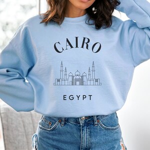 May include: Light blue sweatshirt with the word "CAIRO" arched across the top in black, and "EGYPT" at the bottom. A black line drawing of a cityscape is between the words. The sweatshirt is made of a soft material.