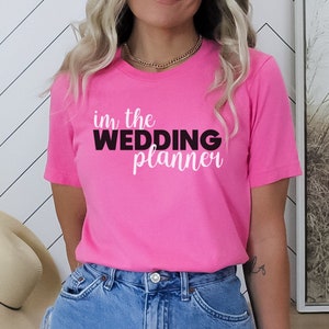 May include: A pink t-shirt with the text "in the wedding planner" in black and white lettering.