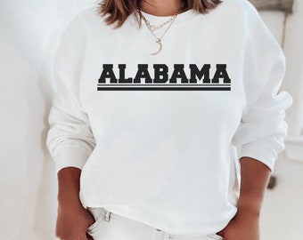 May include: White sweatshirt with black text that reads "ALABAMA" in a bold, blocky font. The text is centered on the sweatshirt and is surrounded by two black horizontal lines.