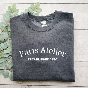 May include: A dark gray sweatshirt with the text "Paris Atelier Established 1954" printed in white.