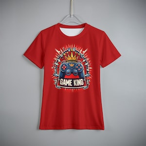 Puede incluir: Camiseta roja con un diseño de un mando de videojuegos azul con una corona dorada, rodeado de líneas blancas. El texto "GAME KING" aparece en una pancarta debajo.