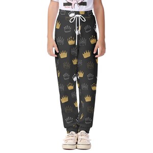 Puede incluir: Pantalones deportivos negros con un estampado repetido de coronas doradas y blancas. Los pantalones tienen cintura con cordón y puños elásticos. El diseño es informal.