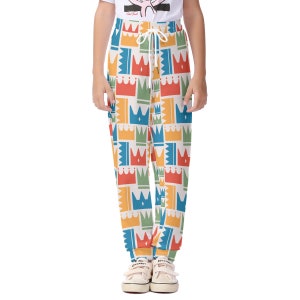 Puede incluir: Pantalones deportivos con un patrón de corona repetido en rojo, azul, verde y amarillo sobre un fondo crema. Los pantalones tienen una cintura con cordón y se usan con zapatillas blancas. El tema general es ropa casual.