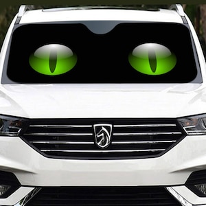 May include: Black car windshield sunshade featuring two large, glossy green cat eyes. The eyes are set against a black background, creating a striking and playful design. The sunshade is designed to fit a car windshield.
