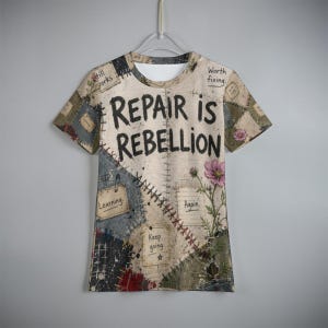 May include: A short-sleeve t-shirt featuring a patchwork design in beige, gray, and blue tones. The text REPAIR IS REBELLION is printed in black across the chest. Additional text includes Worth fixing, Learning, Again, and Keep going.