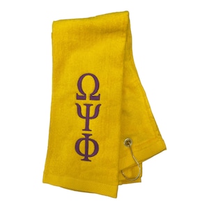 May include: A yellow towel with the Greek letters Omega, Psi, and Phi embroidered in purple. The towel has a loop and a metal hook for hanging. The towel is made of a soft, absorbent material.
