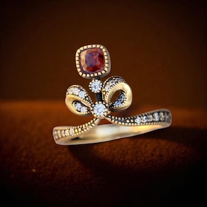Può includere: Un anello color oro con un design a fiocco con piccole pietre chiare scintillanti e una gemma quadrata marrone-rossastra. La fascia dell'anello è anch'essa decorata con piccole pietre chiare.